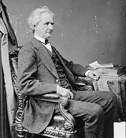 A well-dressed man in middle age facing sideways, sitting in a chair