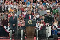 President Trump stands behind a plexiglass shield addressing the crowd. He is flanked by then-Acting Secretary of Defense Mark Esper on the left and Chairman of the Joint Chiefs of Staff Joseph Dunford on the right. A crowd sits on bleachers behind them.
