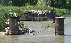 Two vertical cylindrical pilings, joined by horizontal beam near tops, standing in river