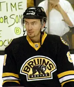 An ice hockey player standing, facing towards the camera. He is wearing a black helmet and a black, yellow and white uniform.