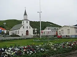 A small white church with a steeply. A small grassy area with flower bushes is in the foreground.