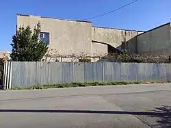 A modern, angular, beige building behind a tall, corrugated metal fence. A small green tree and some vines are visible over the fence.