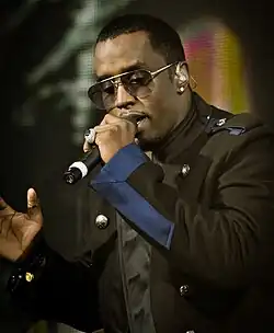 A black man is seen singing into a microphone while wearing a black-blue jacket and sunglasses. A multicolored LED screen is displayed in the background.