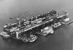 US Navy floating Dry Dock Number 4 in Seeadler Harbor 1945, surrounded by floating barges with workshops and a tugboat, repairing seaplane tender and Navy Liberty ship