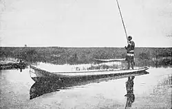 A man holding a cane fishing pole standing in a long flat boat on calm waters