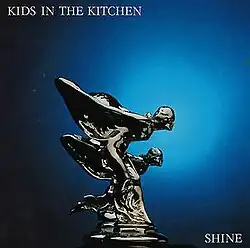 The album's name is seen at the top left, styled in white lettering and all caps over dark blue background. Two winged, female figures are in the middle of the field with a chrome finish. They are striving towards the right as if racing each other. They are mounted on a plinth similar to a car's hood ornament. At the bottom right is the album's name, again in all caps, white lettering.