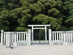 White torii gate behind a concrete fence in front of trees.