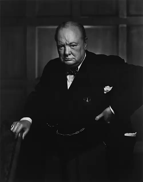 Three-quarter length portrait photograph of Churchill, 67, wearing a suit, standing and holding into the back of a chair