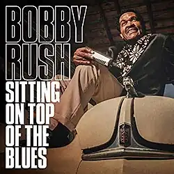 Bobby Rush, sitting on the hood of a vintage car and holding a harmonica