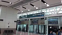 Interior of a modern airport terminal building with glass windows at the top. A sign hanging from the ceiling reads "skytrain Station 3 / D26-D39, E1-E35". Passengers are boarding/alighting a train in the bottom right corner.