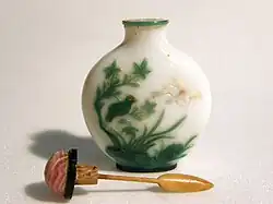 A white and green snuff bottle with plants, a small green bird, and a white flower. Next to the bottle is a brown spoon connected to a coral-colored stopper