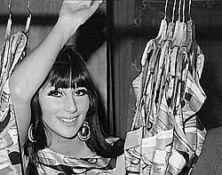 Cher smiles with her arm raised, wearing large hoop earrings and a patterned outfit, surrounded by bold geometric-patterned hanging garments.