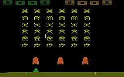 A horizontal rectangular video game screenshot that is a digital representation of a battle between aliens and a laser cannon. The brown aliens hover above three red blocks. Below the blocks is a small green triangular shaped cannon.