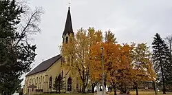 yellowish brick church with a spire partially obscured by autumn leafed trees