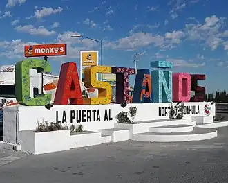 A sign depicting the city name of Castaños. Each letter is a different color. The black text under the letters reads as "la puerta al norte de coahuila", which roughly translates to "the door to the north of Coahuila"