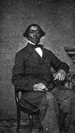 A grainy black-and-white image of a black man with hair projecting over his ears seated in a chair wearing a frock coat and bow tie - from The Autobiography of William Henry Johnson, Argus 1900