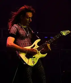 A man wearing black clothing and a chain necklace, holding an electric guitar