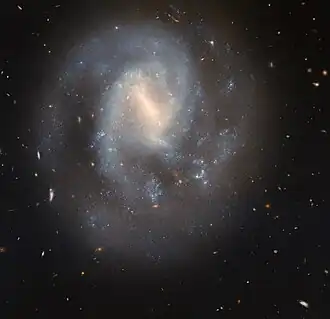 A spiral galaxy with a generally soft and slightly faint appearance. It glows most brightly around the pale yellow bar across its centre. It has two spiral arms which wrap around the centre, quickly broadening out to join a wide, faint circular halo around the galaxy. Glowing, sparkling patches in the disc show stars forming in nebulae. Behind the galaxy, distant galaxies appear as orange dots on a black background.