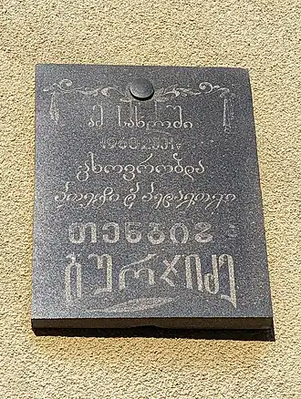 A commemorative plaque using Mkhedruli for the upper four lines and Mtavruli for the lower two, with each line written in a different typeface and showing variant glyphs, like დ with a single curl and ჯ with reduced second stroke. A ligature of და ("and") with ა placed above დ can be seen in the fourth line.
