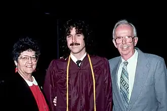 from left to right stands an elderly woman with short, black hair, with glasses and a red dress, an early-middle-aged man with long hair and a maroon-colored academic dress, and an elderly man with short-grey hair and a grey suit and glasses. All three are smiling.