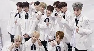 Twelve young men dressed in white suits with black bow ties and silver embellishments posing together in a backstage setting, making hand gestures.