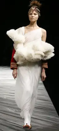 Woman wearing ankle-length slimline ivory gown with large ruffles on the chest and red sleeves, with white tights and red flat shoes