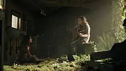 Dina watches as Ellie plays guitar in an abandoned music store overtaken by nature