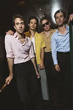A photo depicting the band The Vaccines, taken in a dimly lit room and directly in front of a staircase, with all four members standing together and looking directly into the camera at a slight downward angle.