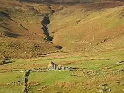 A ruined stone building sits in an empty landscape with a steep slope beyond.