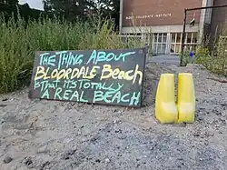 A photograph of the sandy shores of Bloordale Beach, a mannequin butt, and a sign saying The thing about Bloordale Beach is that it is totally a real beach.
