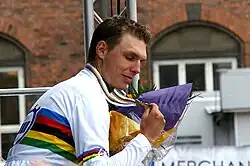 A man in his late twenties wearing a mostly white cycling jersey with rainbow stripes across the midsection and biceps. He has a medal hung around his neck, and clutches it with his right hand. In the other hand is a bouquet of flowers.