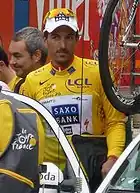 A cyclist in a yellow jersey enters a car with a bicycle on top of it. Other people are visible behind him.
