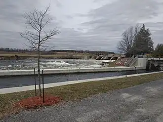 A narrow boating channel is in the foreground, past a small tree. In the background is a dam that is creating rapids in the Ontonabee River. The sky is grey, and the picture was taken in mid-April 2019.