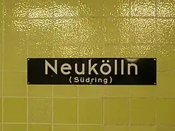 A dark blue sign on a yellow tiled station wall, bearing the name 'Neukölln (Südring)'
