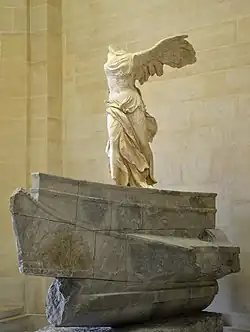 White marble sculpture of a female figure striding forward on a grey marble ships prow, with large wings extending behind her. The statue is missing its original arms and head.