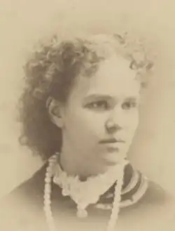 A young white woman with curly hair parted center. She is wearing a high frilly white collar and a strand of large, light-colored beads.