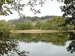 A view across Loch Arthur shows a body of water in the foreground reflecting trees on the other bank. A hill rises in the distance.