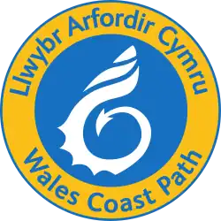A blue circle containing a white shell stylised with a dragon tail enclosed within a yellow ring with the words Wales Coast Path in Welsh and English written on it