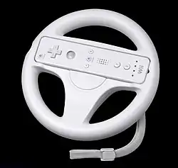 Close up of a white plastic steering wheel. At the center of the wheel is a slot with a Wii Remote, a white handheld video game remote controller.