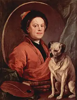A man wearing a red robe and a black hat in a mirror. A small yellow dog with a black nose and ears stands beside the mirror.