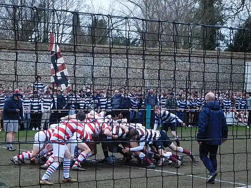 Photograph of a football scrum on a long narrow pitch with ropes and nets along the sides