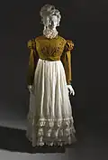 Full-body mannequin wearing a white, floor-length empire waist dress with three rows of ruffles around the bottom and a copper-colored spencer jacket: a long-sleeved, form fitted jacket that reaches the waist and has puffs at the shoulders.