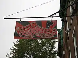 a red and black sign hanging from a building with an image of a shoe and text reading "Wooden Shoe Books: all volunteer anarchist collective"