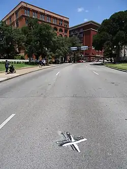 A white cross painted on the middle lane of a three-lane road, view facing towards the Texas School Book Depository