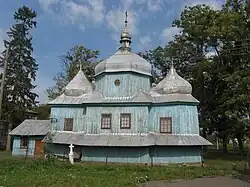 The historic Wooden church of the Holy Mother of God showcases traditional Ukrainian sacral architecture, with its distinctive domes and weathered blue facade standing as a testament to the village’s cultural heritage.