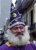 A bearded white man in bright purple and gold robes smiles towards the camera. On his head, a tall pointed purple cap emblazoned with silver and gold stars, moons, and crowns.