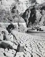 Black and white image of a mud deposit at Hoover Dam. This site can be seen at mile 257, and a member of the survey crew displays a representative sample of one of the mud cubes formed to show the depth of the crevices.