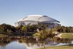 View of AT&T Stadium with a lake and trees in the foreground.